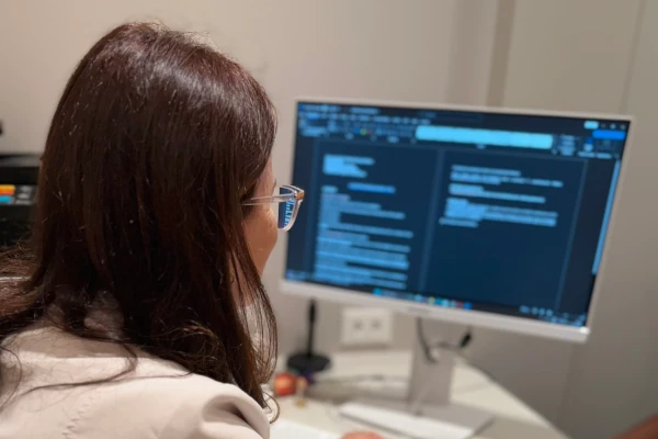 Foto de costas de médica observando tela do computador desfocada ao fundo, com prontuário de paciente aberto na tela.